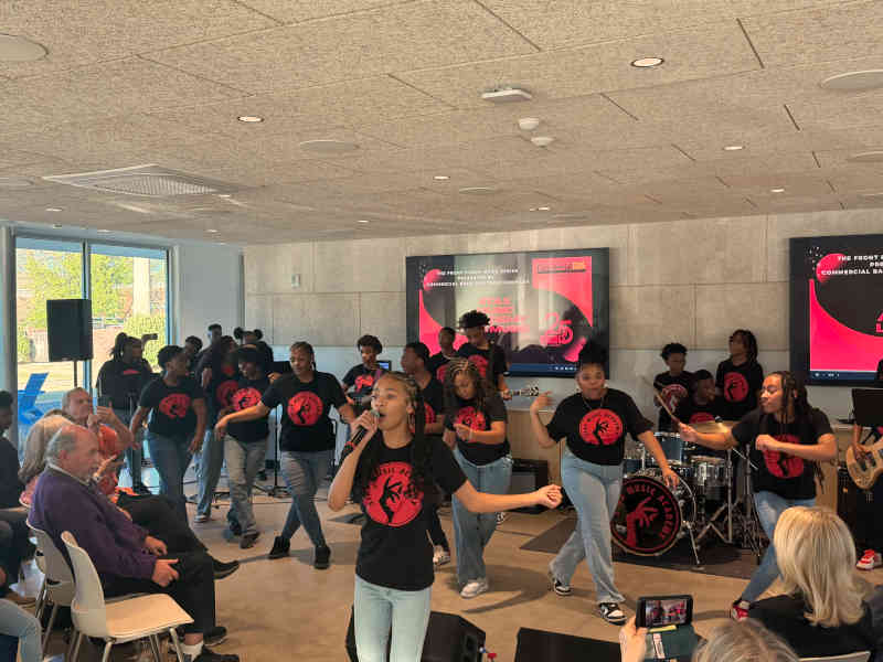 A Stax Music Academy student sings into a handheld microphone at the front of the room while the ensemble performs behind her, an audience seated close by on both sides