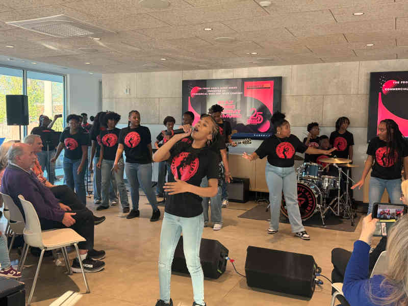 A vocalist reaches for a high note mid-song, band and backing singers behind her beneath screens reading The Front Porch Music Series presented by Commercial Bank and Trust