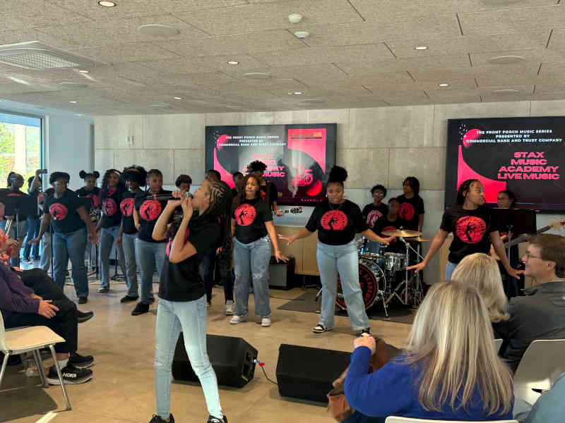 Students in matching Stax Music Academy shirts perform together, with a drum kit and screens reading Stax Music Academy Live Music behind them