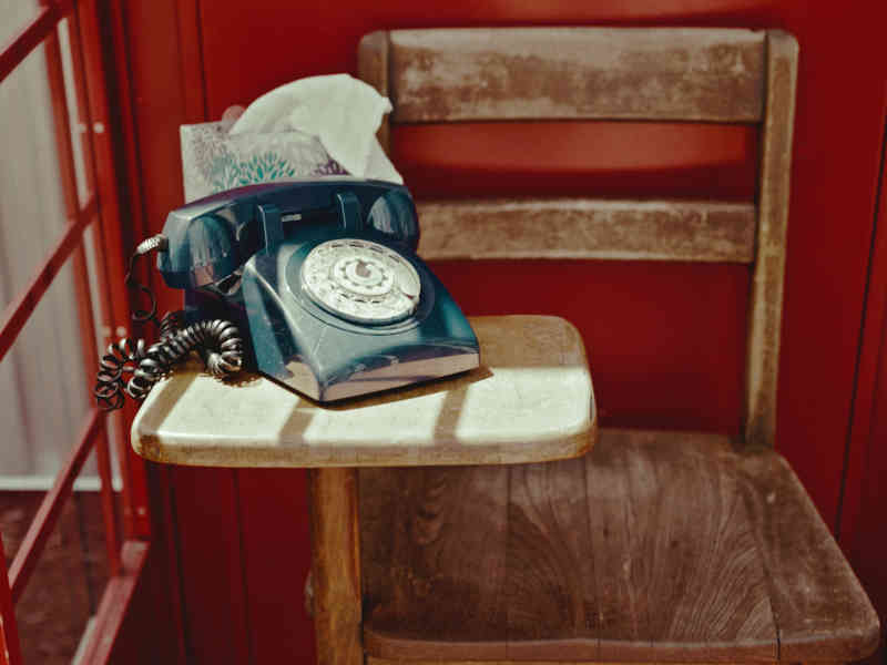 A blue rotary telephone resting on a worn wooden chair inside the red booth, with a box of tissues beside it