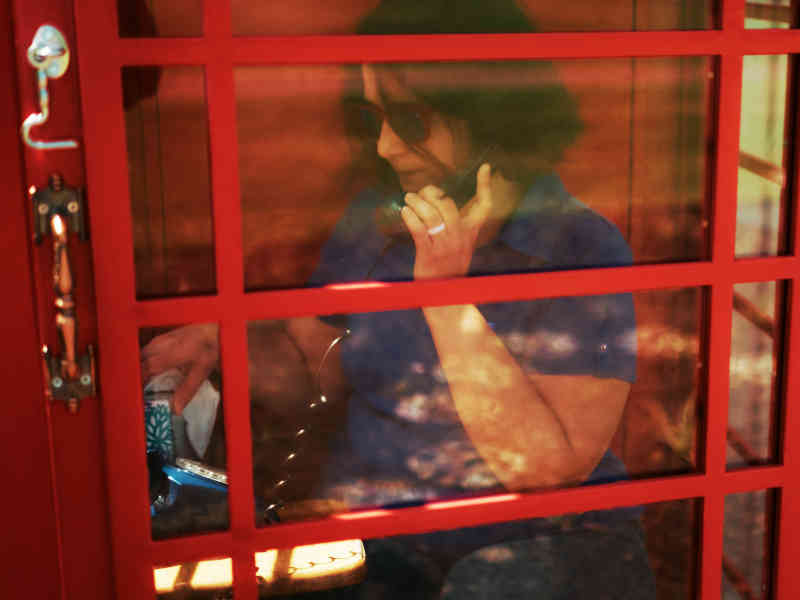 Seen through the glass panes of the booth door, a woman holds the receiver to her ear, head lowered